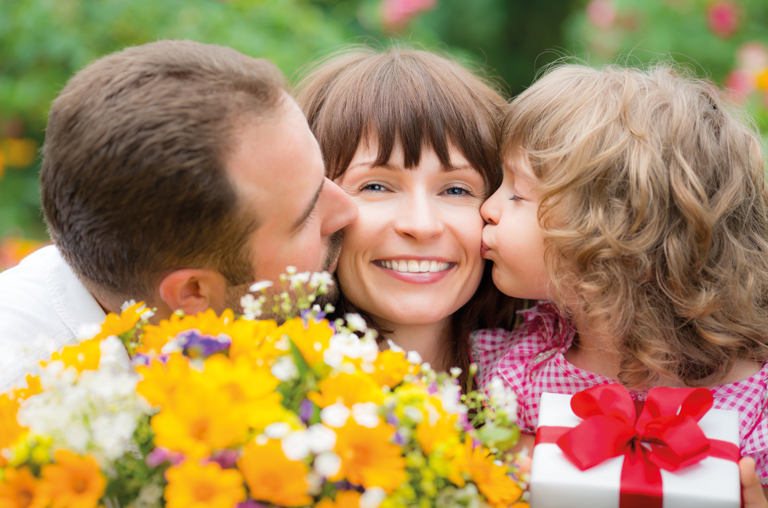 Ein Kind und ein Ehemann küssen die Mama zum Muttertag beide auf die Wange und schenken ihr einen Blumenstrauß